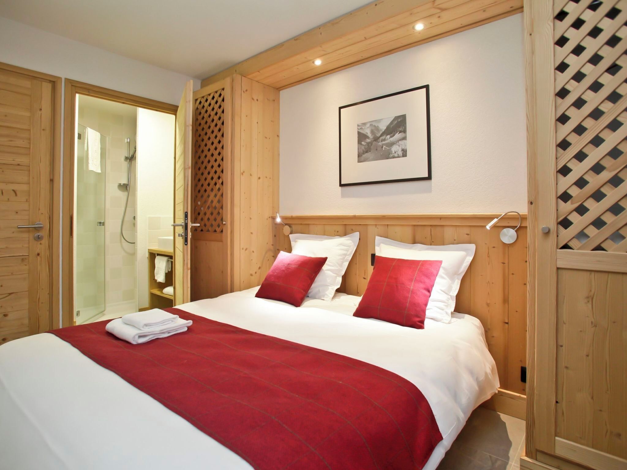 Une chambre d’hôtel avec une tête de lit en bois, un lit avec une literie blanche et rouge, un tableau encadré au mur et une porte en bois avec une paroi de douche en verre.