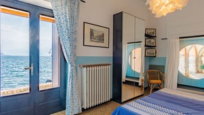 Un dormitori amb vista al mar, un llit amb llençols blaus i blancs estampats, una cadira de fusta, un radiador i una finestra gran amb cortines.