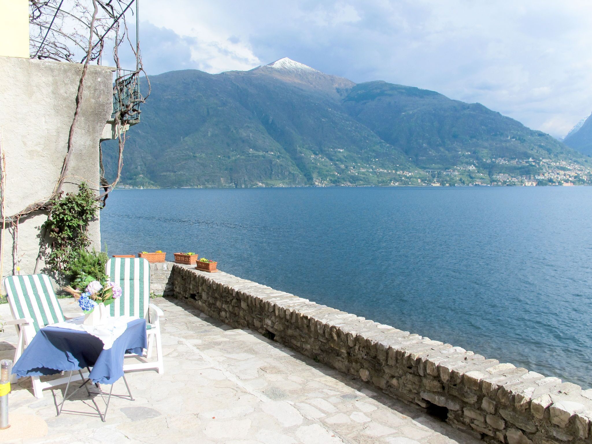 A terrace with stone flooring, two striped lounge chairs, and a table with a blue tablecloth. A stone wall borders the terrace, and a mountain with a snow-capped peak is visible in the background.