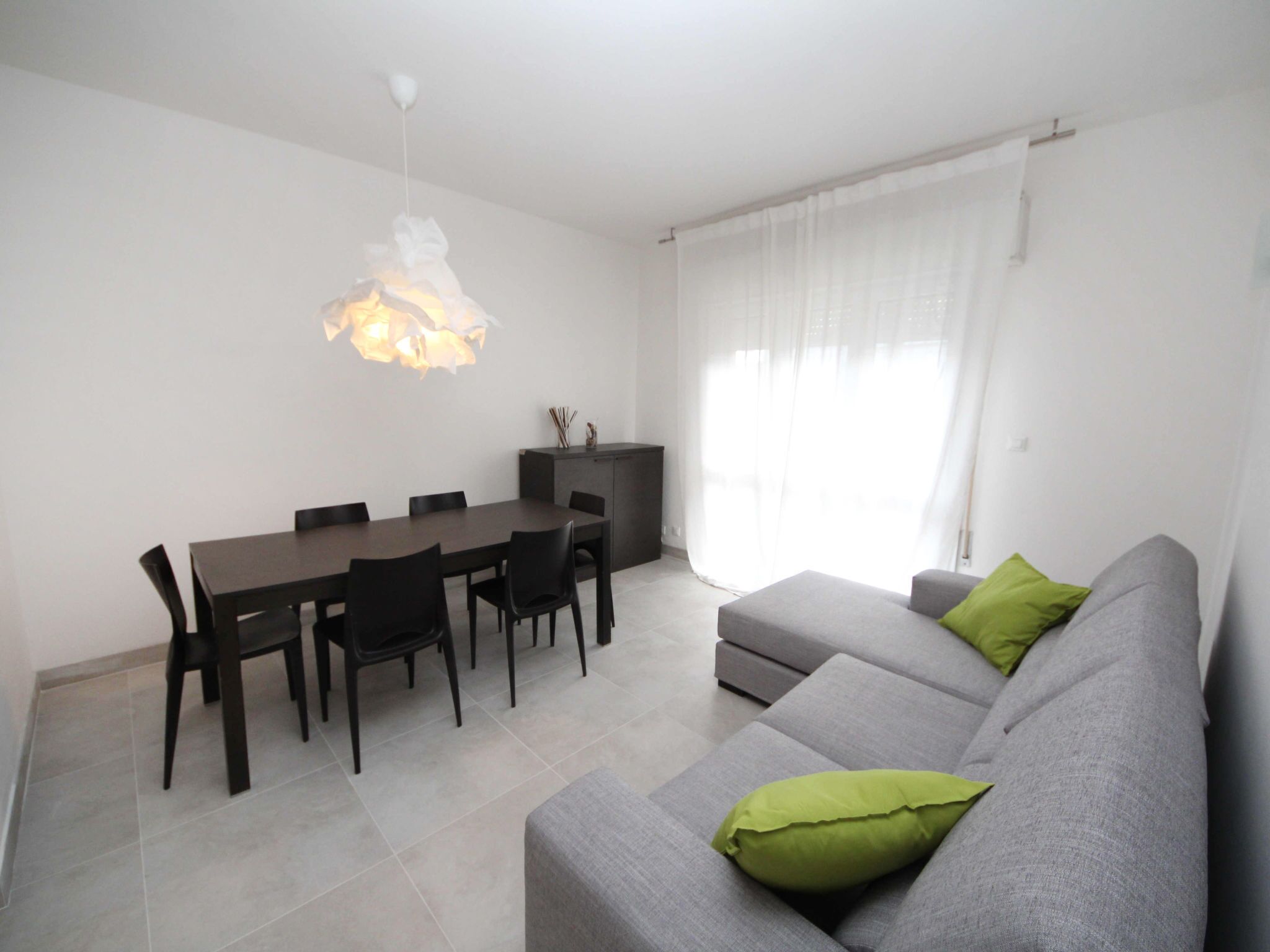 Een moderne woonkamer met een grijze bank, een eettafel met stoelen en een hanglamp.