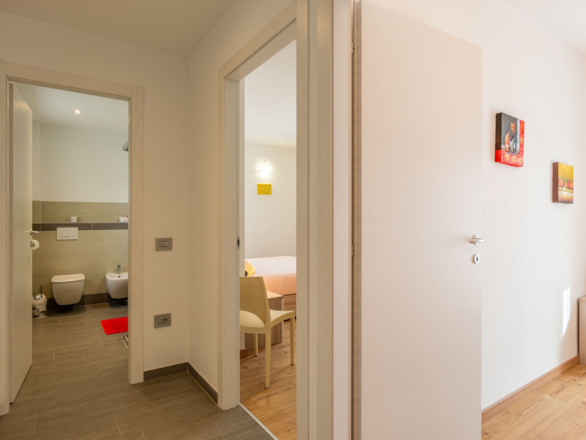 Habitación de hotel con piso de madera, una cama con manta roja, una silla y una mesita. Cuenta con un baño con inodoro y lavamanos.
