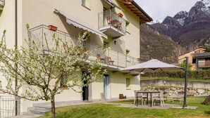 Ein zweistöckiges Gebäude mit Balkonen, einer Terrasse und Gartenmöbeln, vor einer Bergkulisse.