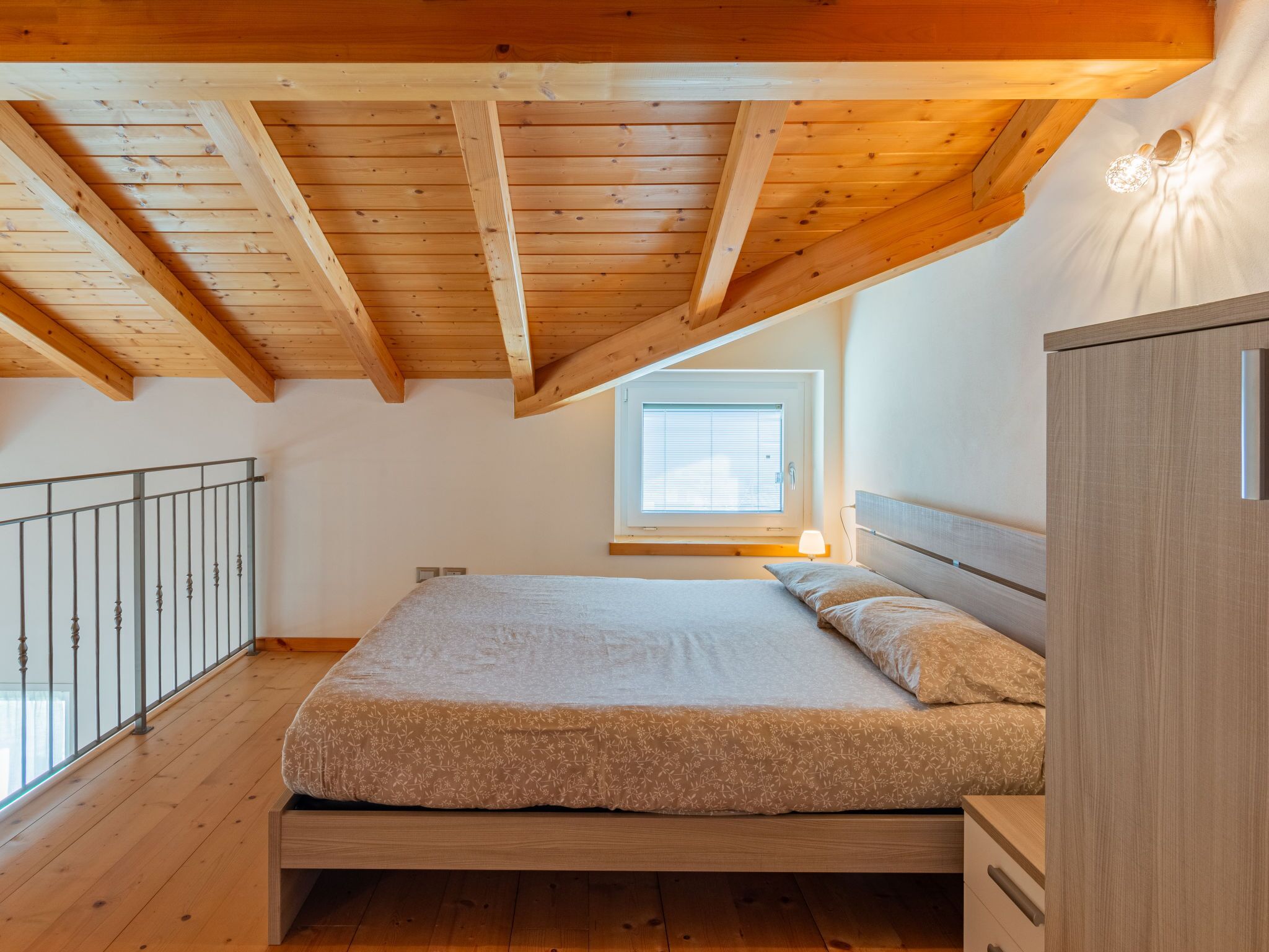 Un dormitorio con techo de madera, una cama con colcha estampada, una mesita de noche y una ventana con persianas.