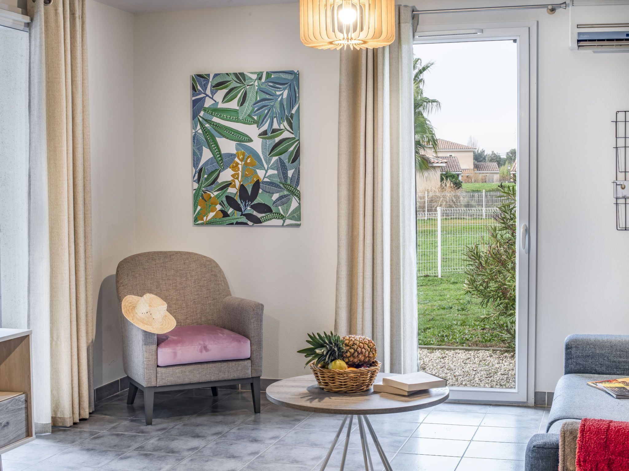Ein Zimmer mit grauem Sessel, rundem Tisch mit Obstkorb, Zimmerpflanze und Blick auf einen Garten durch eine Schiebetür.
