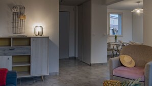 Ein modernes Wohnzimmer mit einer Couch, einem Couchtisch mit Buch und Obstkorb, einer Wandleuchte und einem Essbereich im Hintergrund.