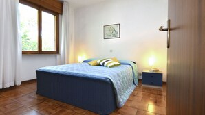 A bedroom with a bed, a window with curtains, a bedside table with a lamp, and a wall-mounted picture. - Missana by Interhome (Lignano Riviera)