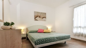 A bedroom with a bed, bedside tables, a lamp, a painting on the wall, and a window with curtains. - Villaggio Laura by Interhome (Lignano Riviera)