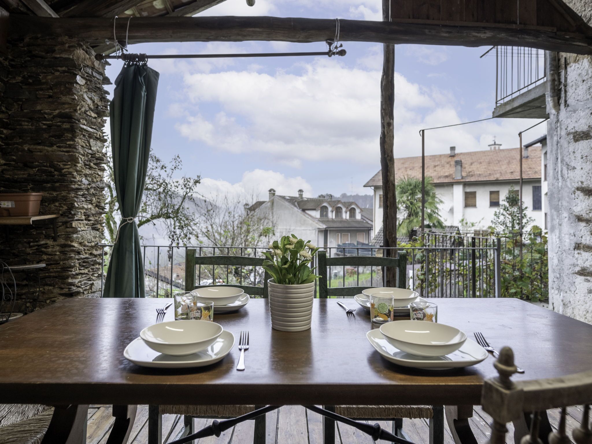 An outdoor dining area with a rustic wooden table set for two, surrounded by stone walls and a view of buildings in the background.