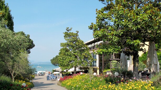 A pathway lined with colorful flowers leading to a building with outdoor seating and a view of the sea.