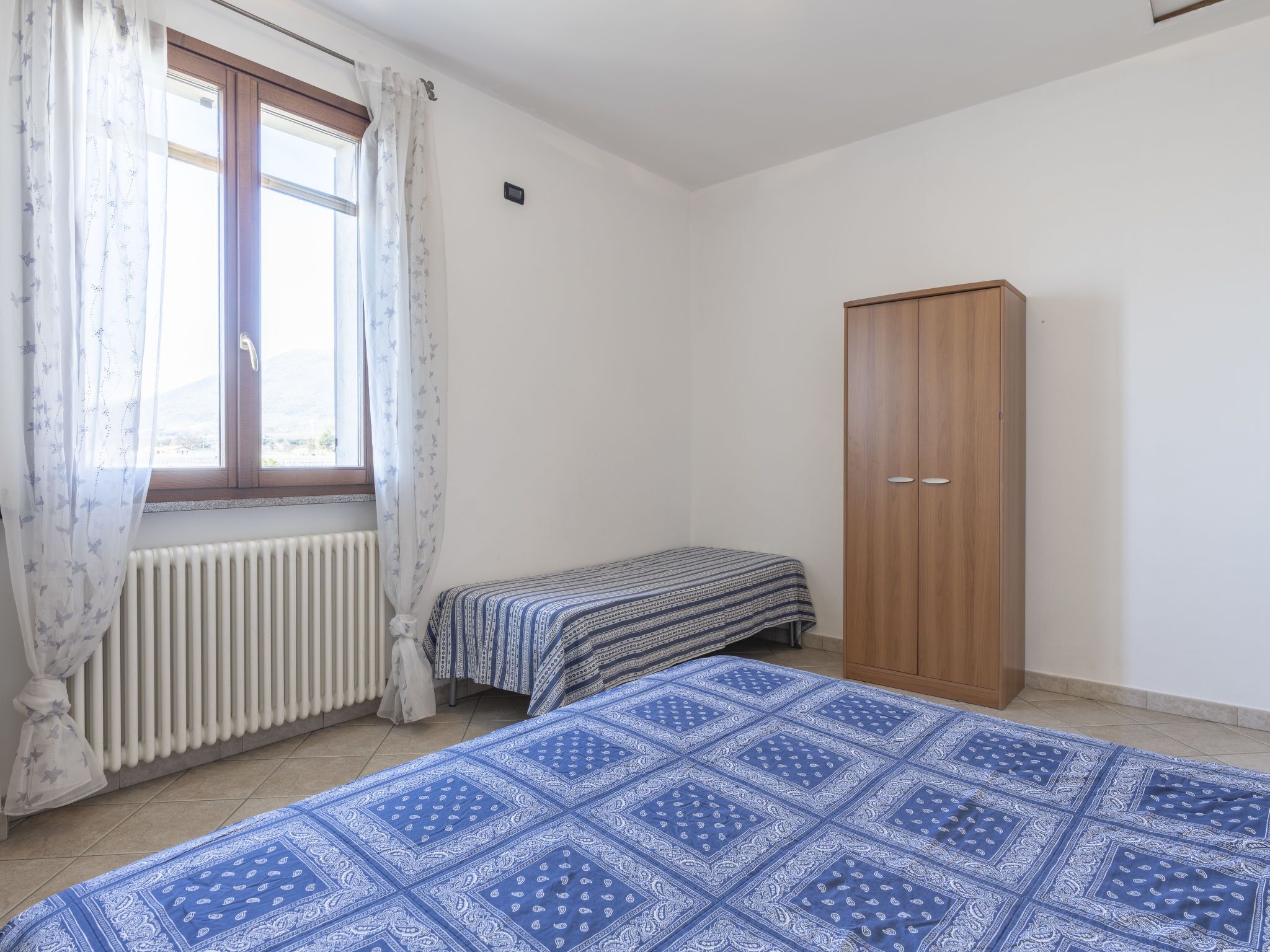 Une chambre avec une fenêtre, un radiateur, deux lits et une armoire.