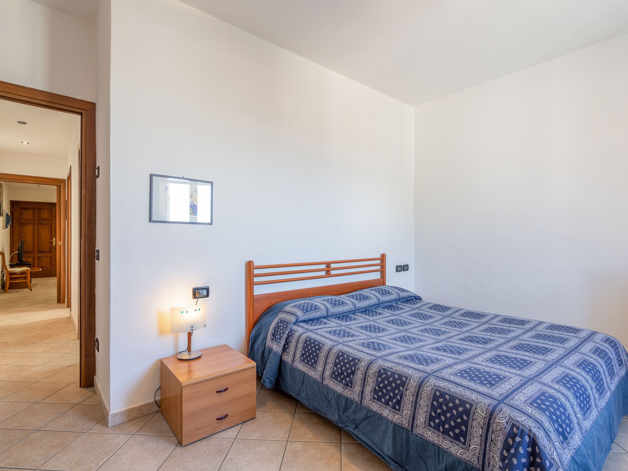 A bedroom with a wooden bed, a bedside table with a lamp, and a door leading to another room.
