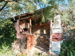 A rustic outdoor kitchen with stone walls and a wooden roof, surrounded by greenery. - Glicine by Interhome (Vendone)