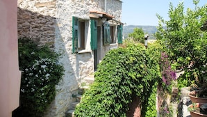 Un bâtiment en pierre avec des volets verts, un jardin où poussent différentes espèces de plantes et une vue sur le paysage.