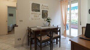 Un coin repas avec une table et des chaises, un mur sur lequel figure une date et un lieu, une fenêtre avec des rideaux et un téléviseur posé sur un meuble.