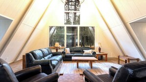 A spacious loft with a high ceiling, large windows offering a forest view, leather sofas, and a wooden coffee table. - Charming Modern A-frame Chalet with Hot Tub--8 min walk to Country Club (Angel Fire)