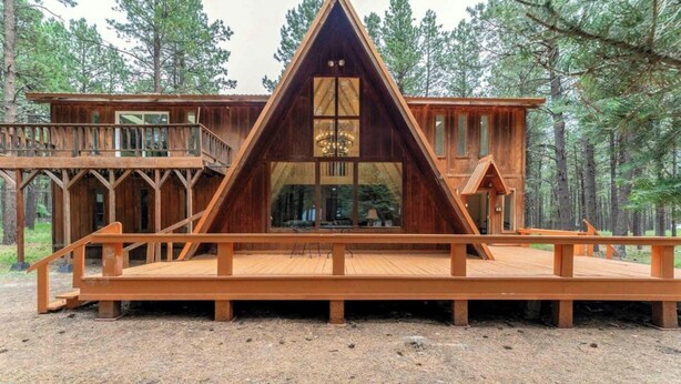 A wooden cabin with a triangular roof, large windows, and a deck. - Charming Modern A-frame Chalet with Hot Tub--8 min walk to Country Club (Angel Fire)