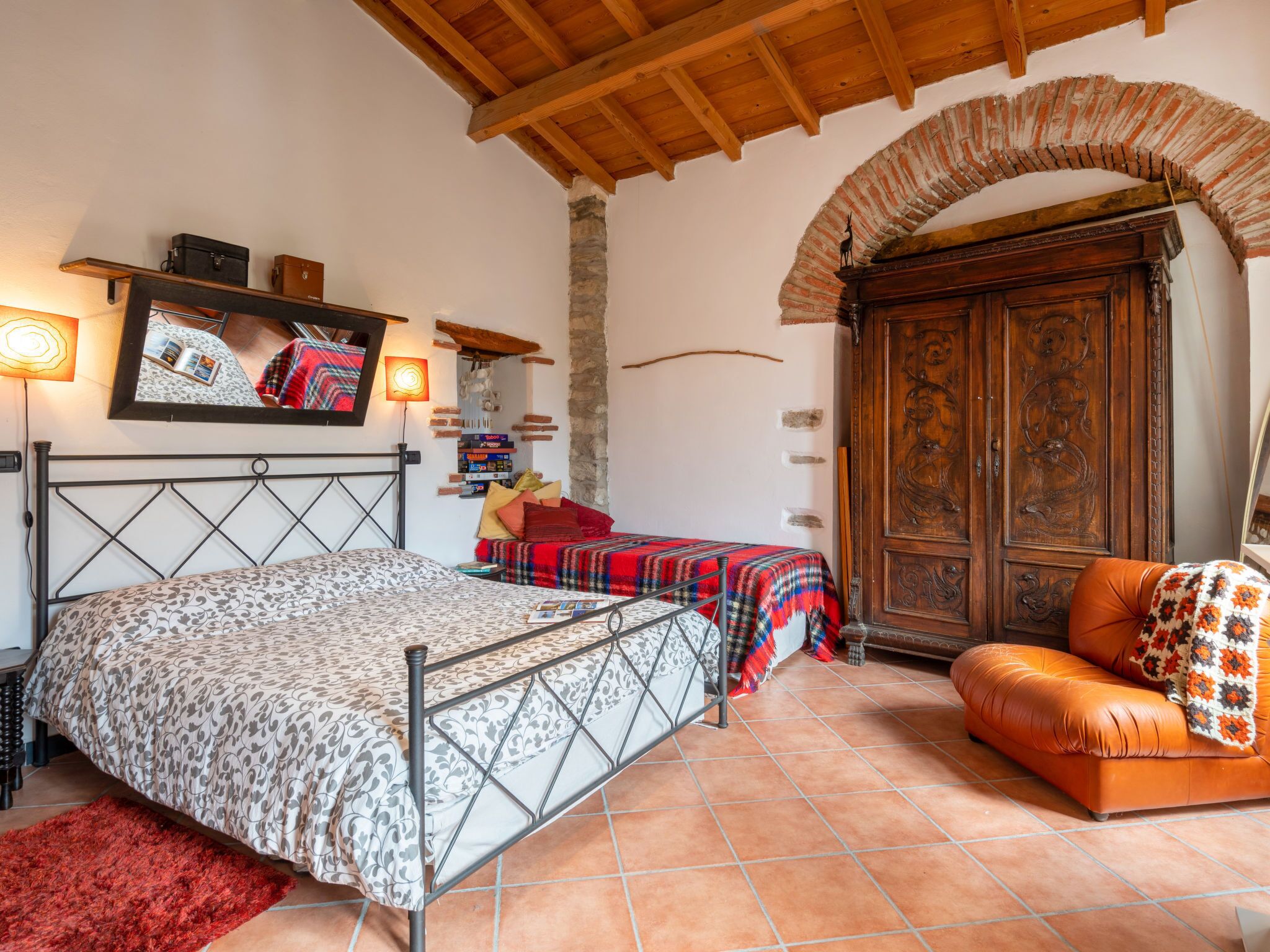 A bedroom with a bed, a red rug, a wooden arched doorway, a leather sofa, and a stone wall.