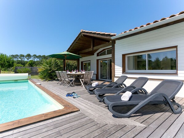Area piscina con solarium in legno, sedie a sdraio, tavolo e sedie, e un grande ombrellone.