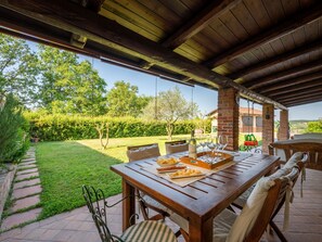 An outdoor dining area with a wooden table and chairs, surrounded by a brick wall and a view of a garden and trees.