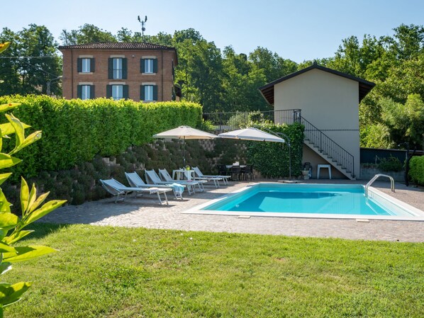 A swimming pool with lounge chairs and umbrellas, surrounded by greenery and a building with a tiled roof.