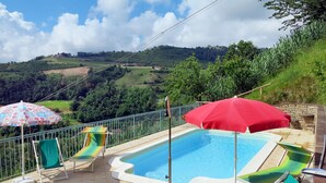 Una piscina amb gandules i ombrelles, envoltada per una tanca metàl·lica, amb vistes a un paisatge amb monts.
