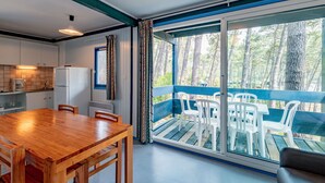 A modern kitchen with a wooden table and chairs, a refrigerator, and a window overlooking a balcony with white plastic chairs.