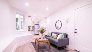 A modern living room with a grey sofa, a wooden coffee table, and a vase of flowers. A kitchen is visible in the background.