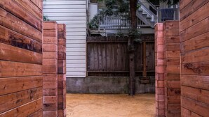 A narrow wooden gate leading to a sandy courtyard with a wooden fence and a building in the background.