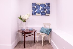 Ein Eckzimmer mit weißer Wand, Holzboden, einem kleinen runden Tisch mit zwei Schalen, einem weißen Stuhl mit blauem Kissen und einer Vase mit weißen Blumen.