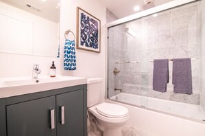 Un baño moderno con lavamanos blanco, muebles oscuros, inodoro y zona de ducha con mampara de vidrio.