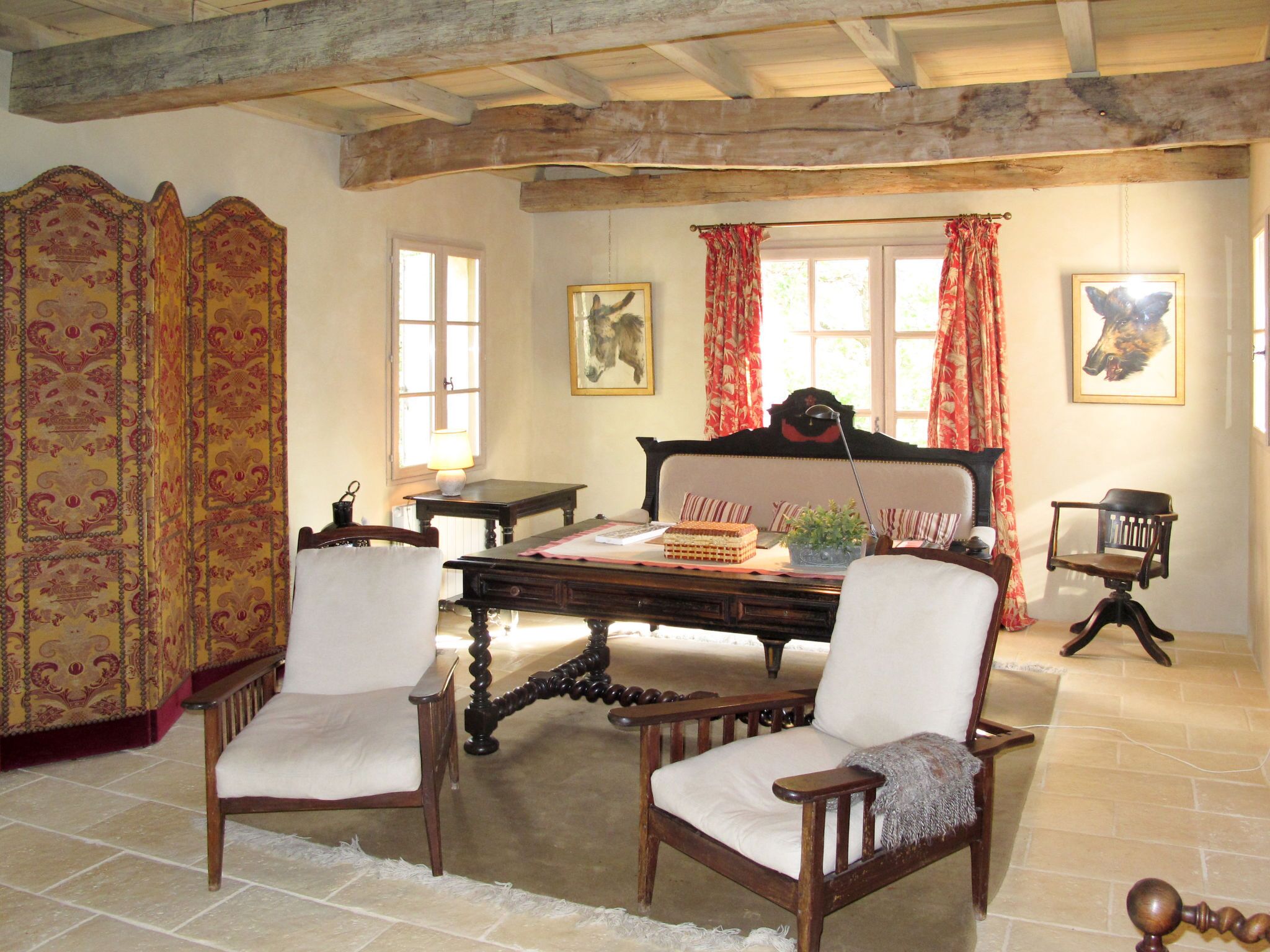 A traditional room with a wooden ceiling, a large bed with a headboard, two upholstered chairs, a wooden table with a lamp, and a window with curtains.