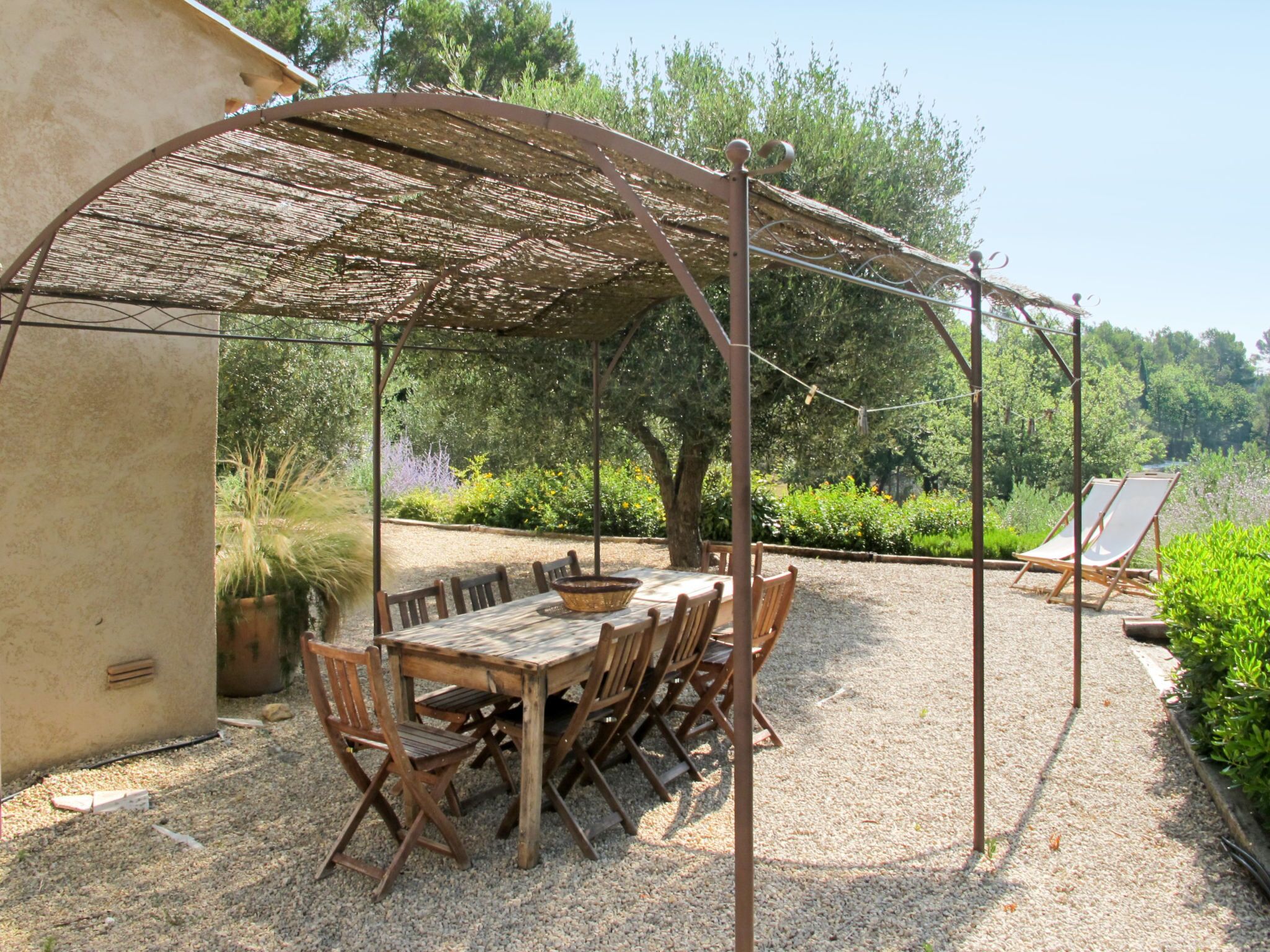 Une terrasse extérieure avec une table et des chaises en bois, une structure d’ombrage en fibres tressées et un sol en gravier.