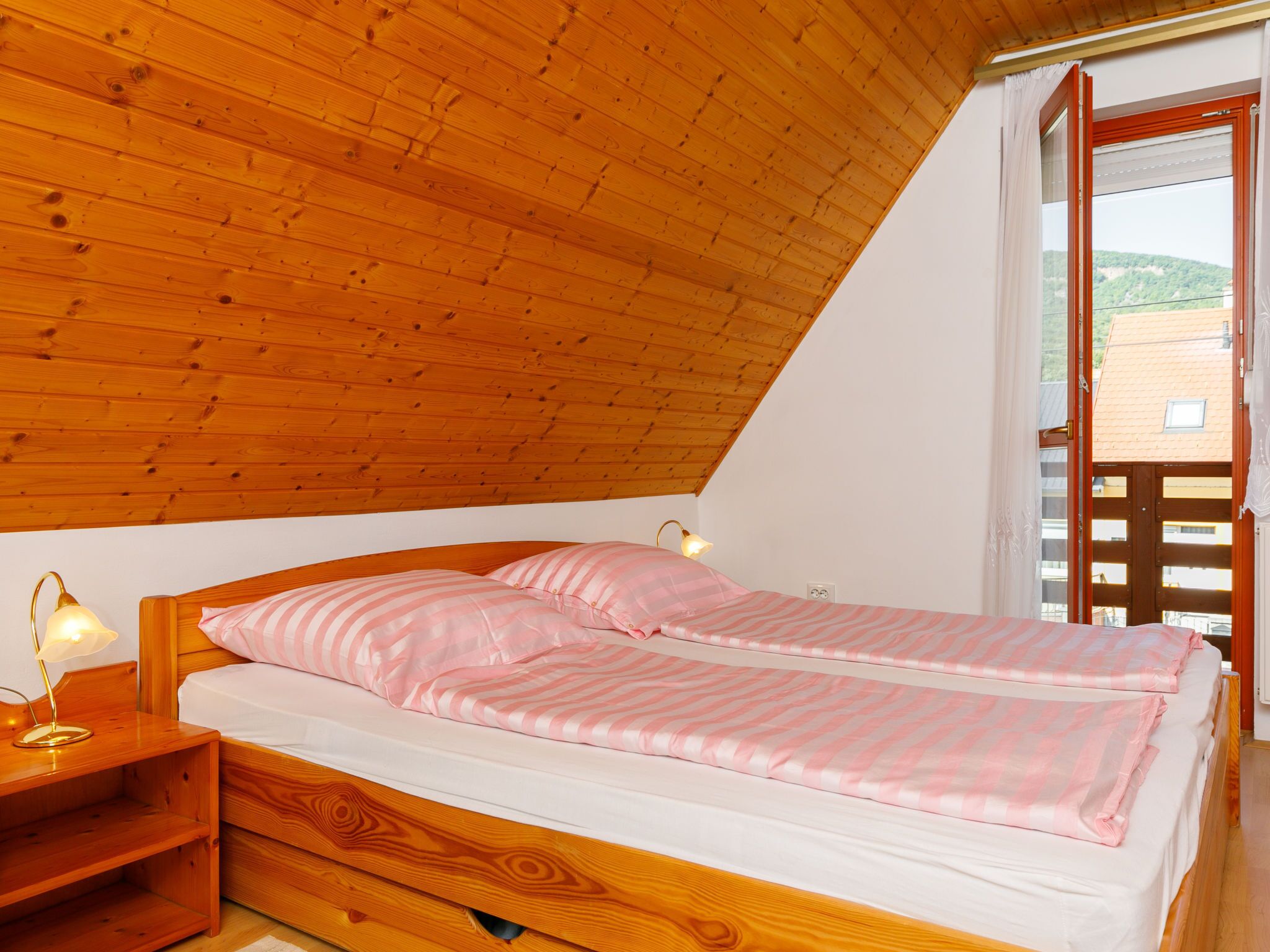 Ein Schlafzimmer mit Holzbalkendecke, einem Bett mit rosa-weiß gestreifter Bettwäsche, einem Nachttisch mit Lampe und einem Fenster mit Blick auf Gebäude.