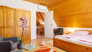 Een slaapkamer met een houten plafond, een bed met roze beddengoed, een bureau met stoel en een zichtbare eethoek door een geopende deur.