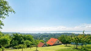 Uma casa com telhado vermelho numa colina gramada com vista para uma cidade e um corpo de água.
