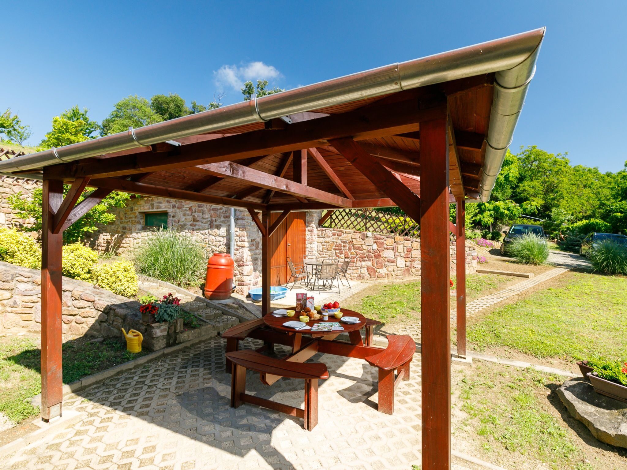 Une pergola en bois, agrémentée d’une table et de bancs, entourée d’un mur de pierre et de verdure.