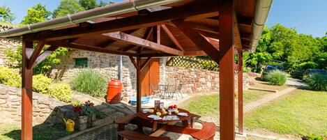 Une pergola en bois, agrémentée d’une table et de bancs, entourée d’un mur de pierre et de verdure.