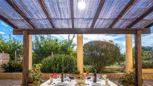 An outdoor dining area with a pergola, two black chairs, a table set with plates, glasses, and cutlery, surrounded by potted plants and a view of a tree and greenery. - Finca Can Corritx by Interhome (Muro)