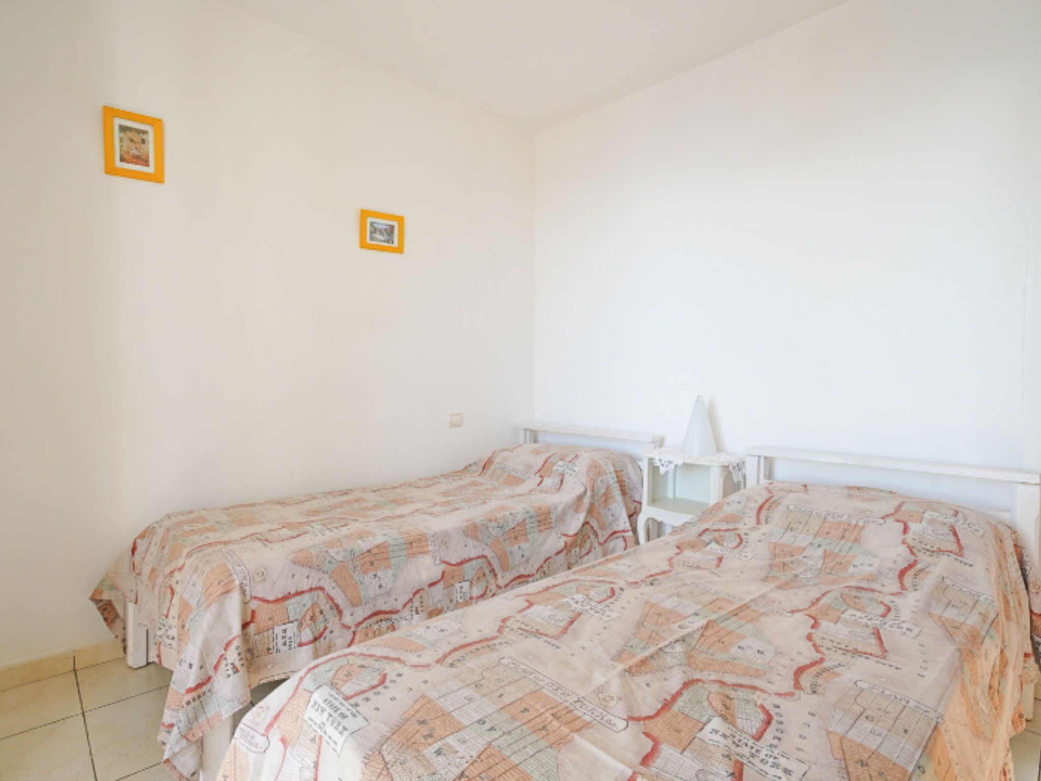 A room with two beds, plain white walls, and two small framed pictures.