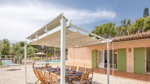 An outdoor dining area with a covered table and chairs, a pool, and a building with green doors. - Mas des Chênes by Interhome (Grimaud)