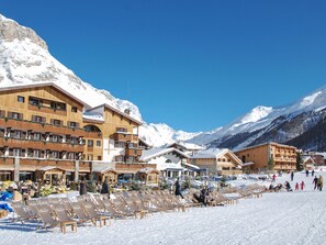 Un hôtel à étages, doté d’une terrasse et dont l’arrière-plan est dominé par une piste de ski.