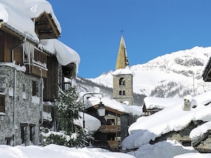 Un pueblo cubierto de nieve con edificios de piedra tradicionales, una torre de iglesia y montañas al fondo.