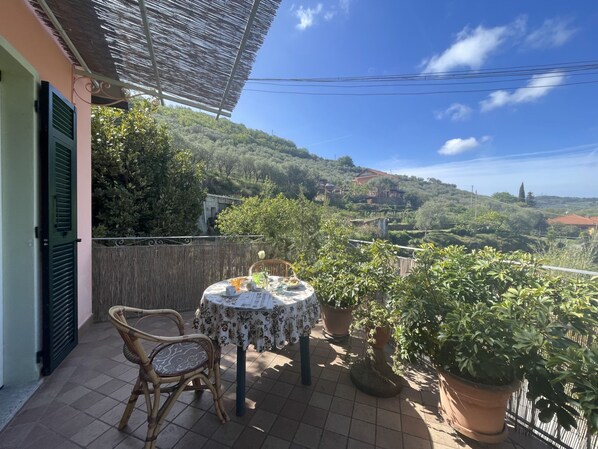 Relax on this serene outdoor space with a round table and two wicker chairs, surrounded by lush greenery and a hilly landscape.