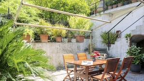 An outdoor patio with a wooden table and chairs, surrounded by potted plants and a stone wall. - Degli Aranci (IMP385) by Interhome (Imperia)