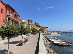 A coastal promenade with a row of colorful buildings, a paved walkway with benches, and boats docked at the pier.
