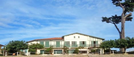 Un edificio de dos plantas con techo plano, contraventanas verdes y un muro de piedra al frente.