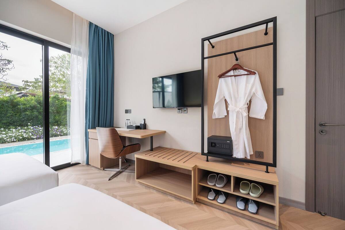 Una habitación de hotel moderna con una cama grande, un escritorio con una silla, un televisor montado en la pared, una bata colgada en un perchero y un mueble para guardar zapatos.