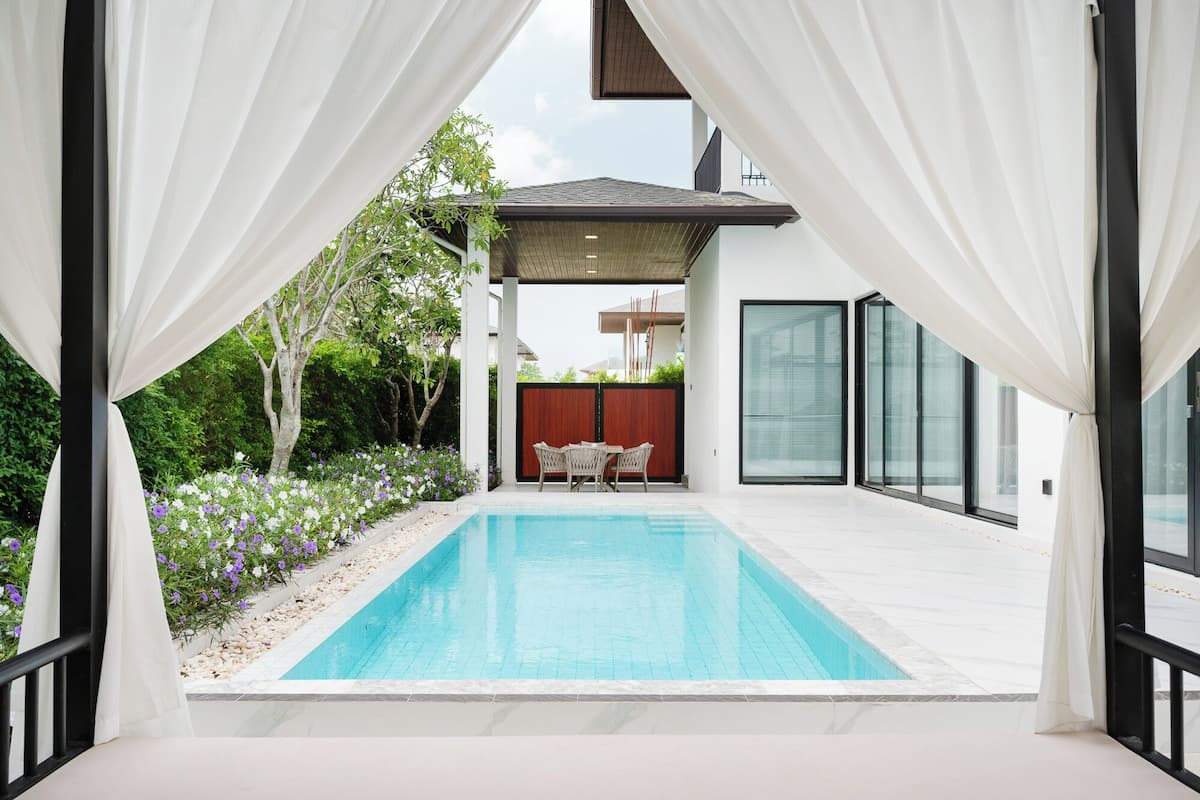 Un área de piscina moderna con piscina azul cristalina, rodeada de cortinas blancas, una valla de madera y una zona de estar con sillas.
