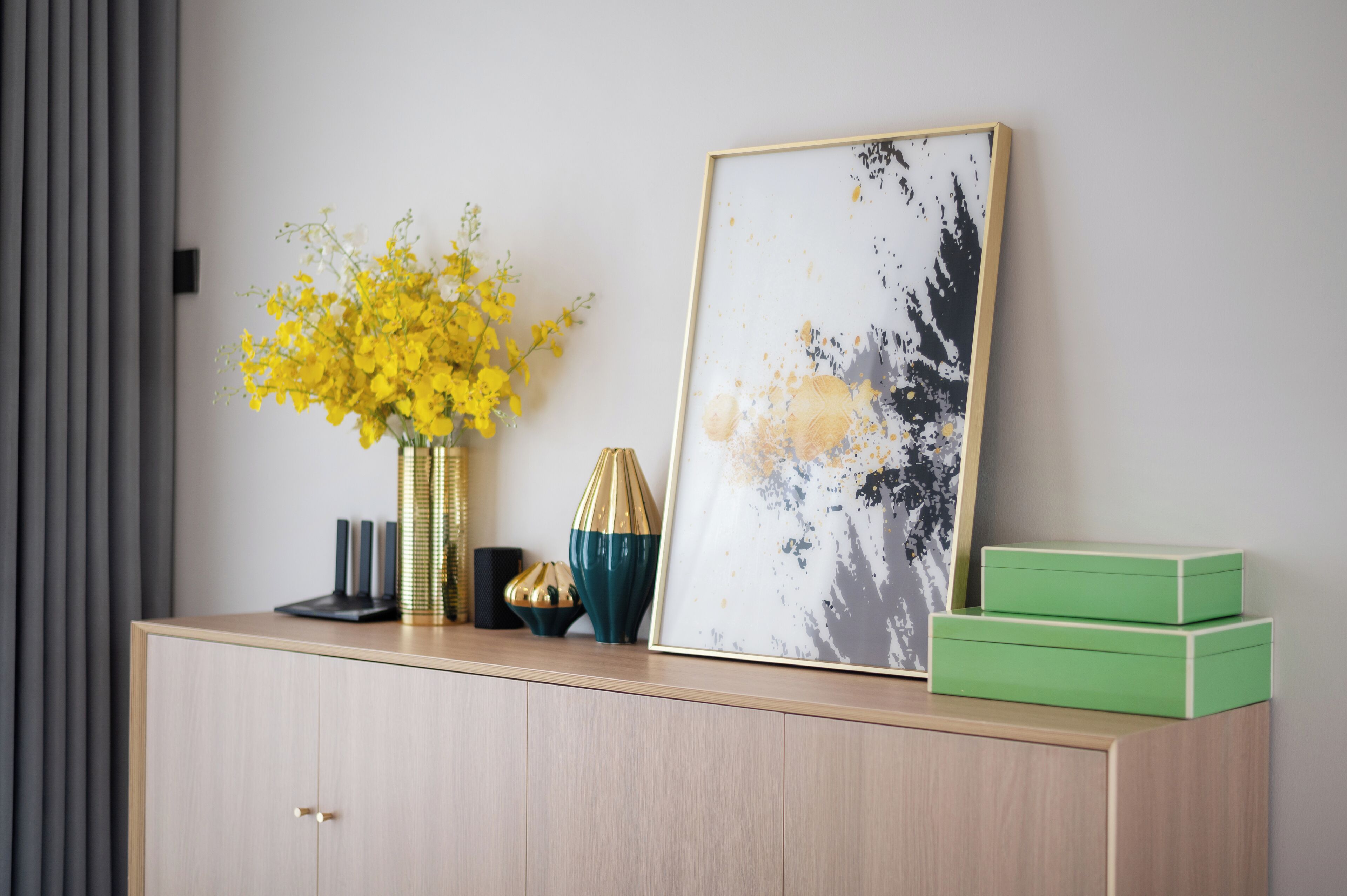 Une pièce au décor minimaliste, avec une armoire en bois, un tableau abstrait encadré, un vase de fleurs jaunes et une boîte verte.