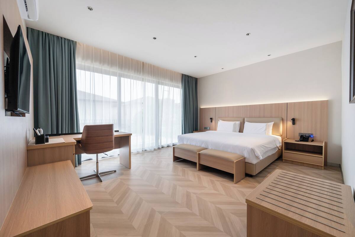 Una habitación de hotel moderna con piso de madera, una cama con cabecera de madera, un escritorio con silla, un televisor montado en la pared y una ventana con cortinas.